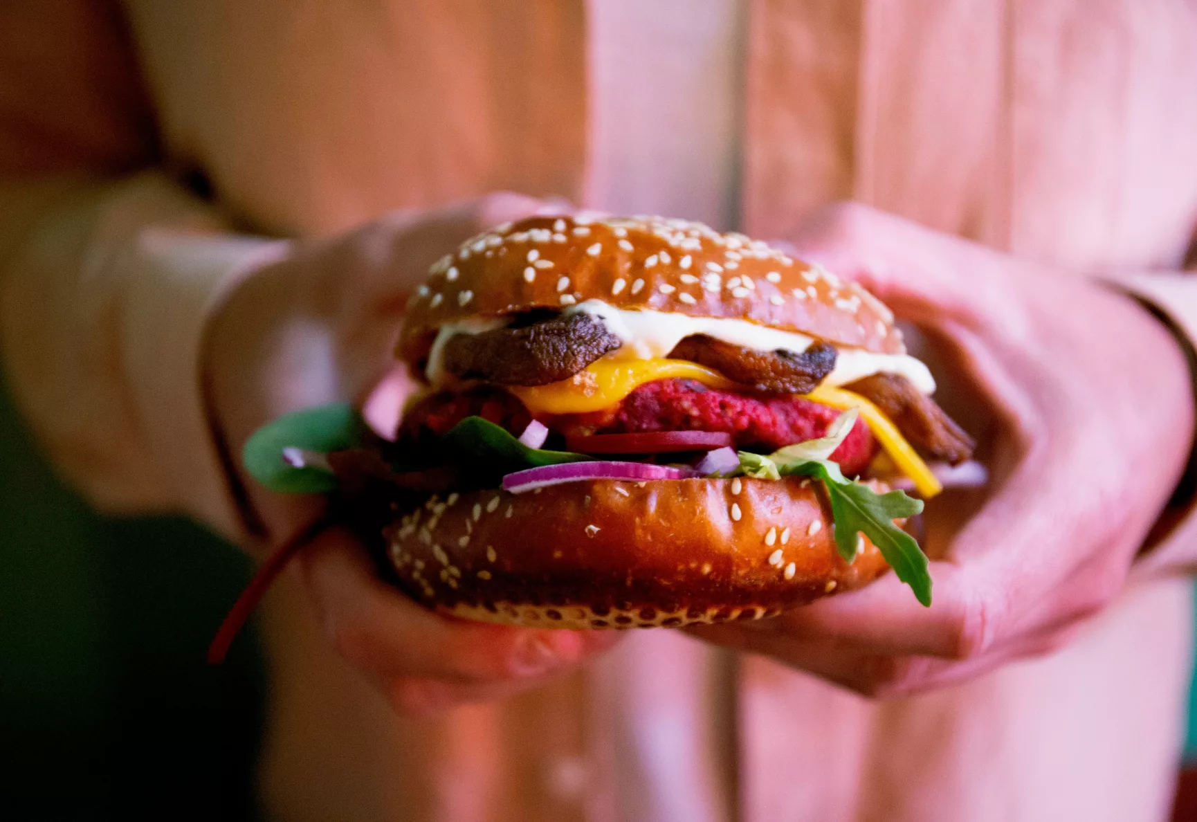 Closeup of someone holding a veggie burger with beetroot, mushrooms, onions and rocket, in a dark seeded bun