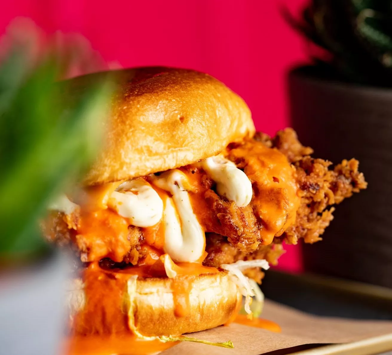 A closeup of a fried chicken burger on brown waxed paper, in a brioche bun with mayo and hot sauce.