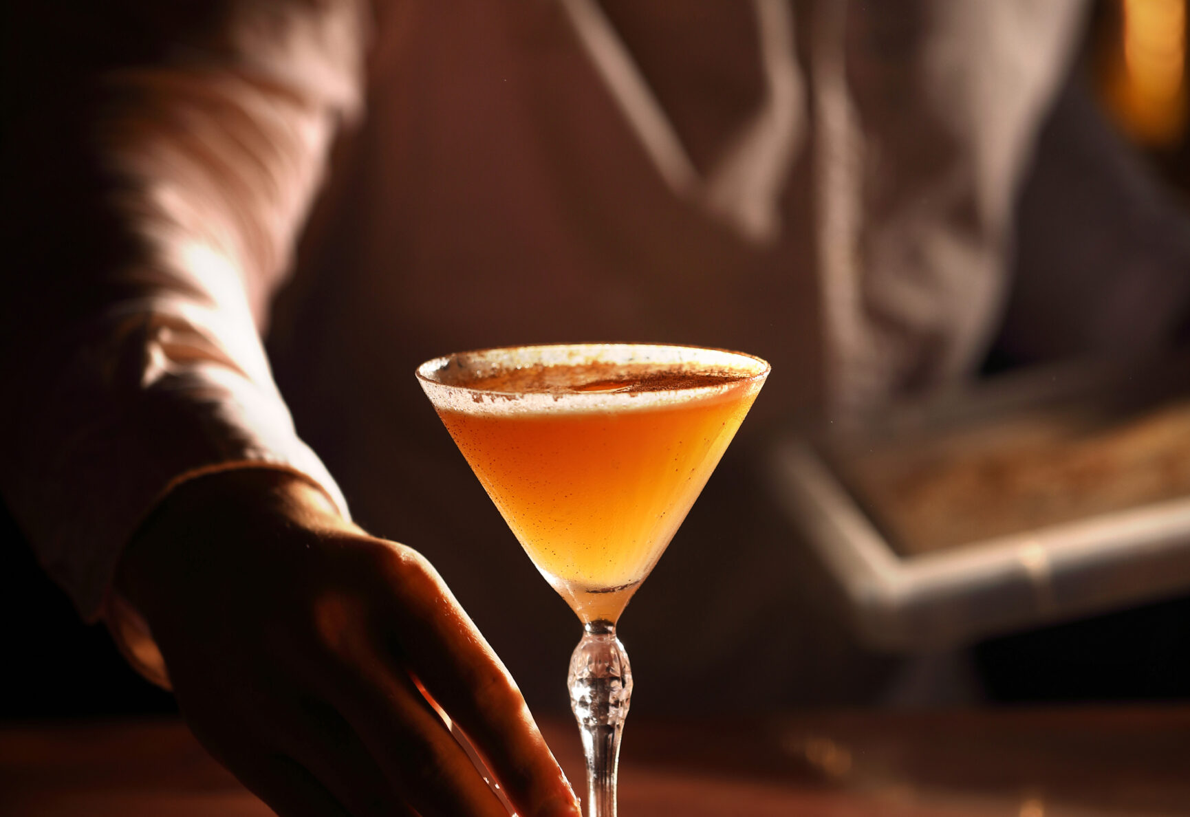 A cocktail in a martini glass being placed by a bartender in a white shirt.