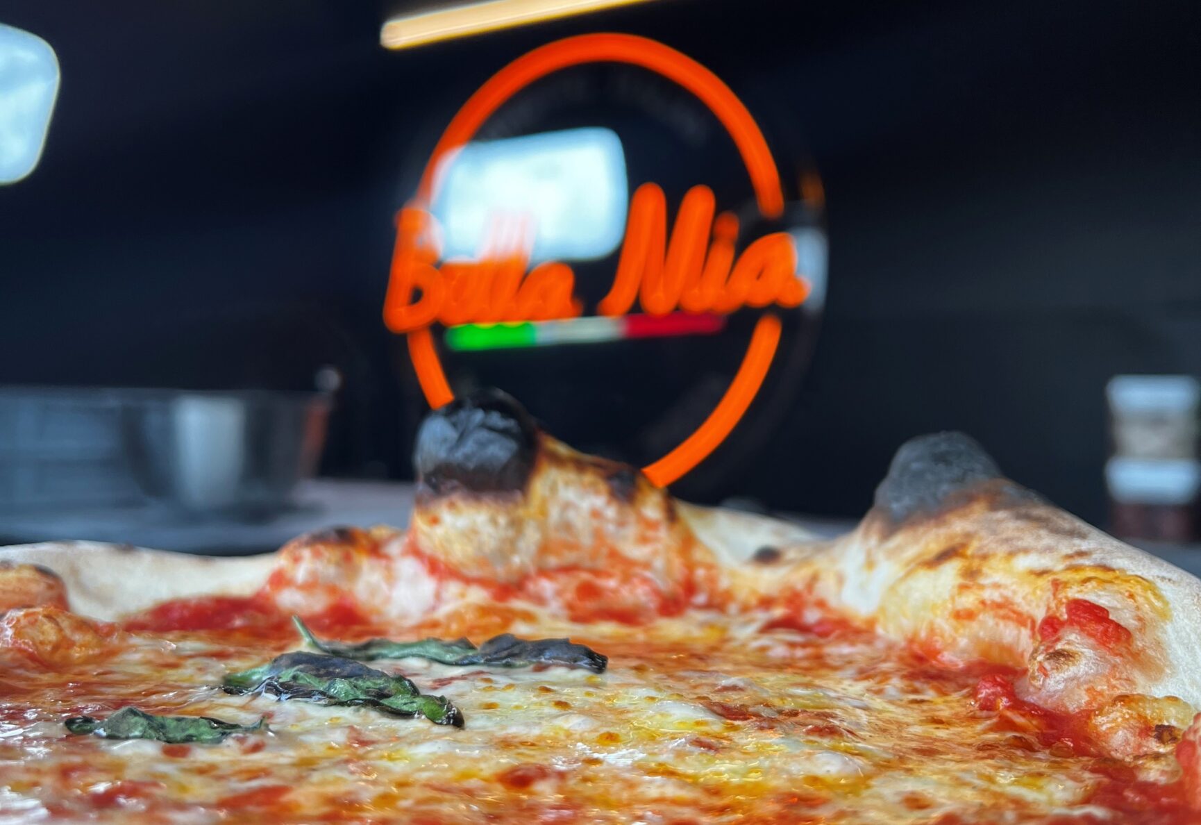 A close up view of a thick crusted, margherita pizza, with the 'Bella Mia' logo in the background.