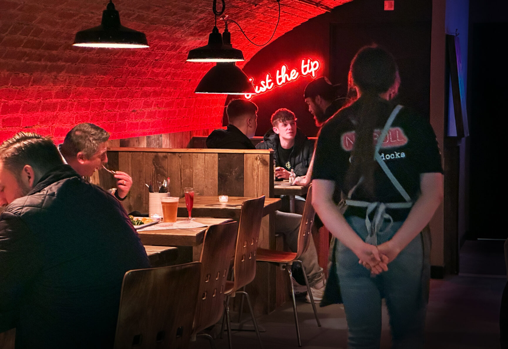 A serving walks with their hands behind their back past table filled with people enjoying food.