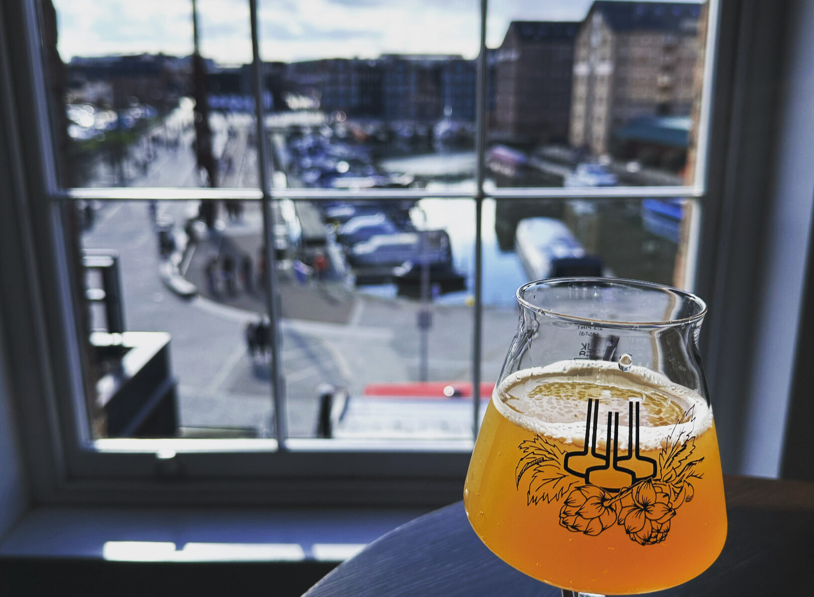 A beer glass sits on a wooden table, with a view of Gloucester Docks seen behind through a sash window.