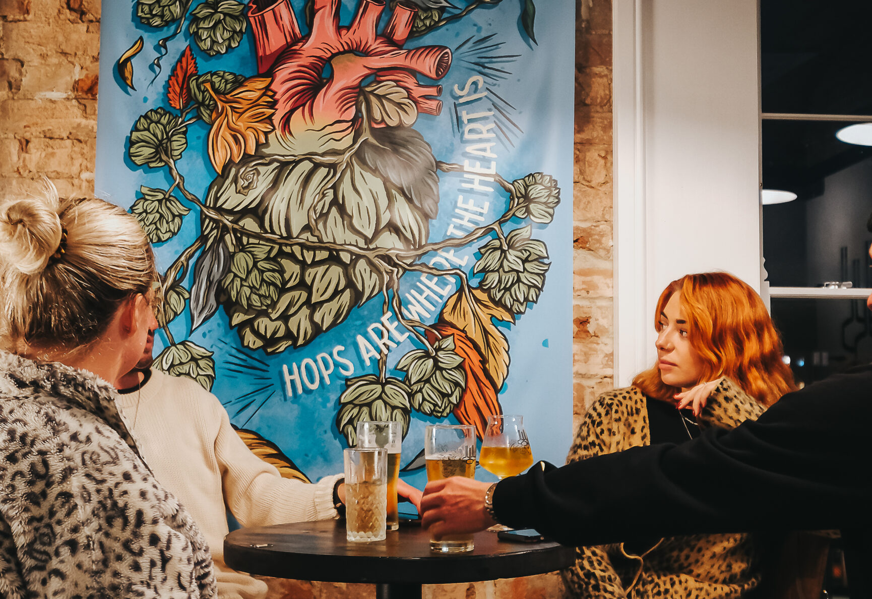 Three friends sat a tall table. A poster with an illustration of the Hop Kettle Logo hangs on the brick wall behind them.