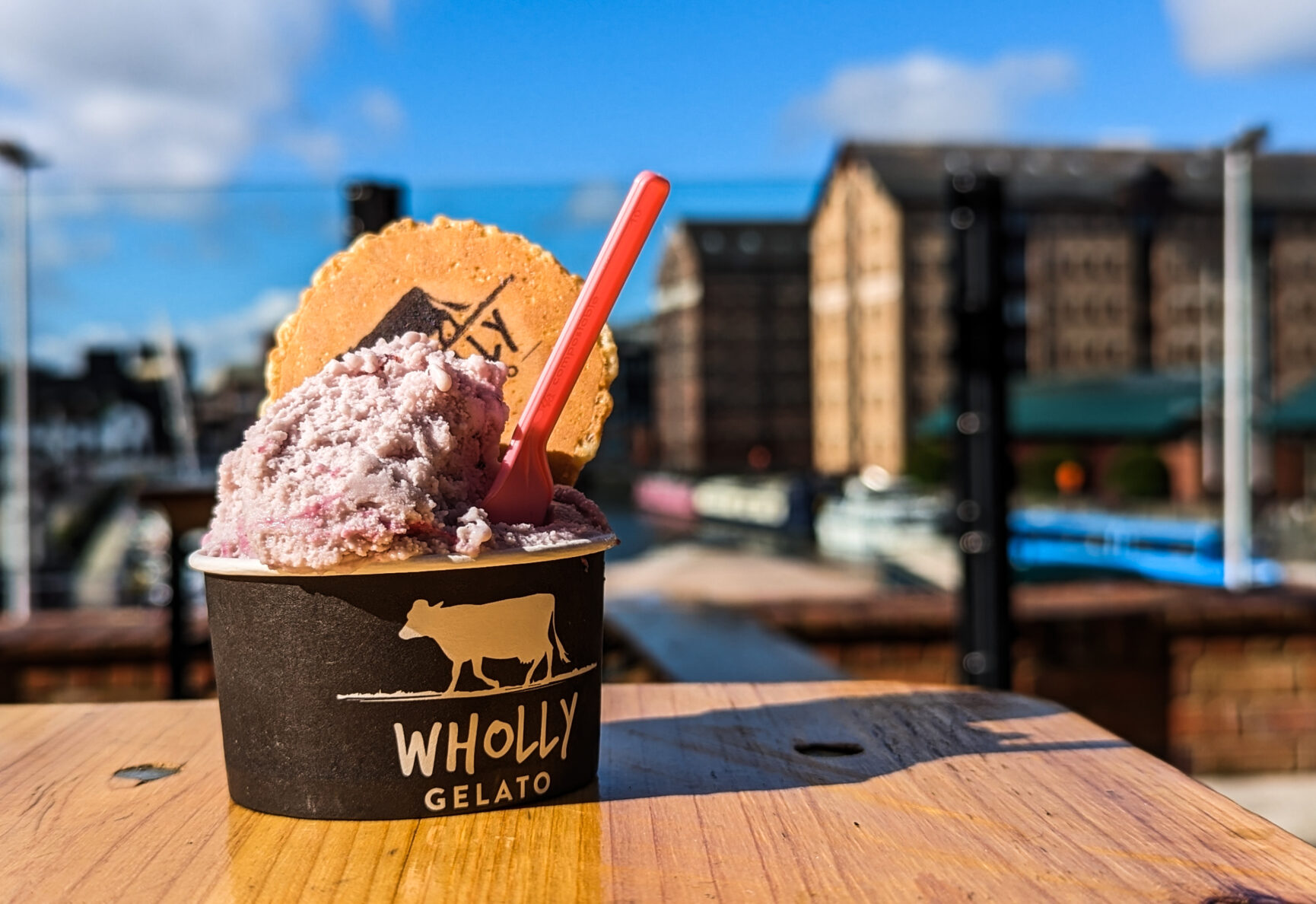 A pot of mixed berry gelato with a spoon and wafer sits on a table on the Food Dock deck with a view of blue sky and the historic Gloucester docks buildings behind.