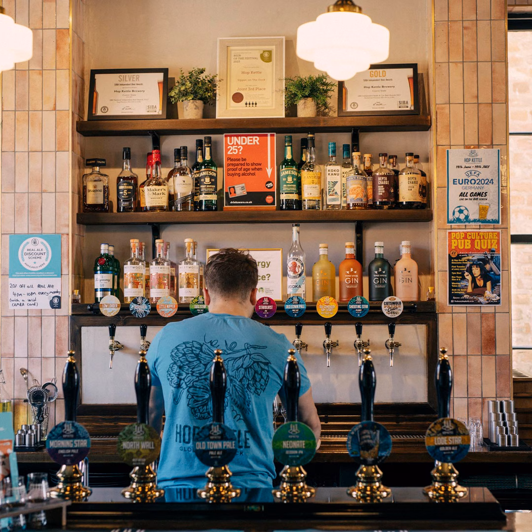 A bartender behind a row of indie beer taps. A selection of spirits can be seen shelving behind the bar.