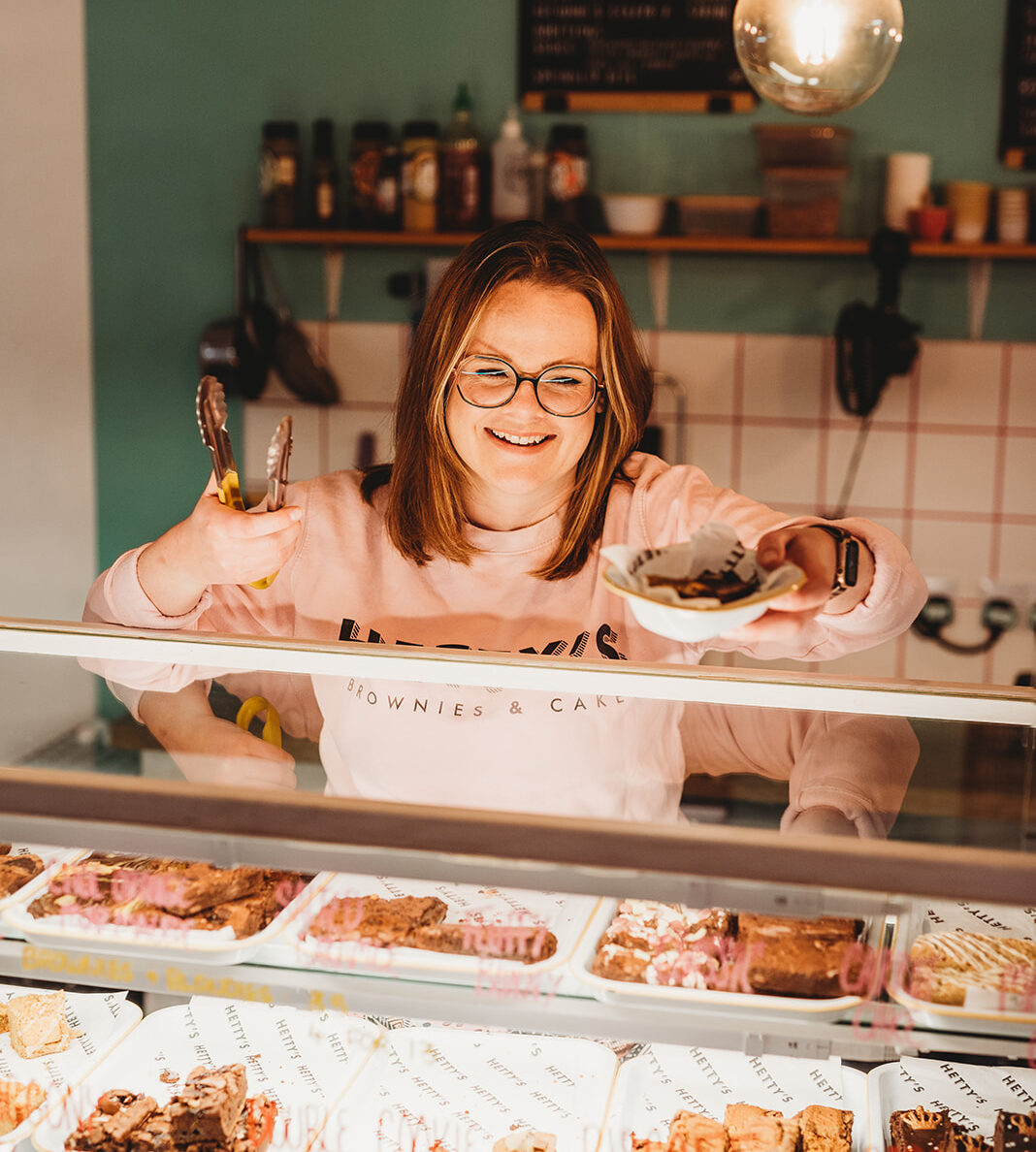 A smiling Hetty placing a plate of brownies on a counter top, with a selection of brownies in a display case underneath.