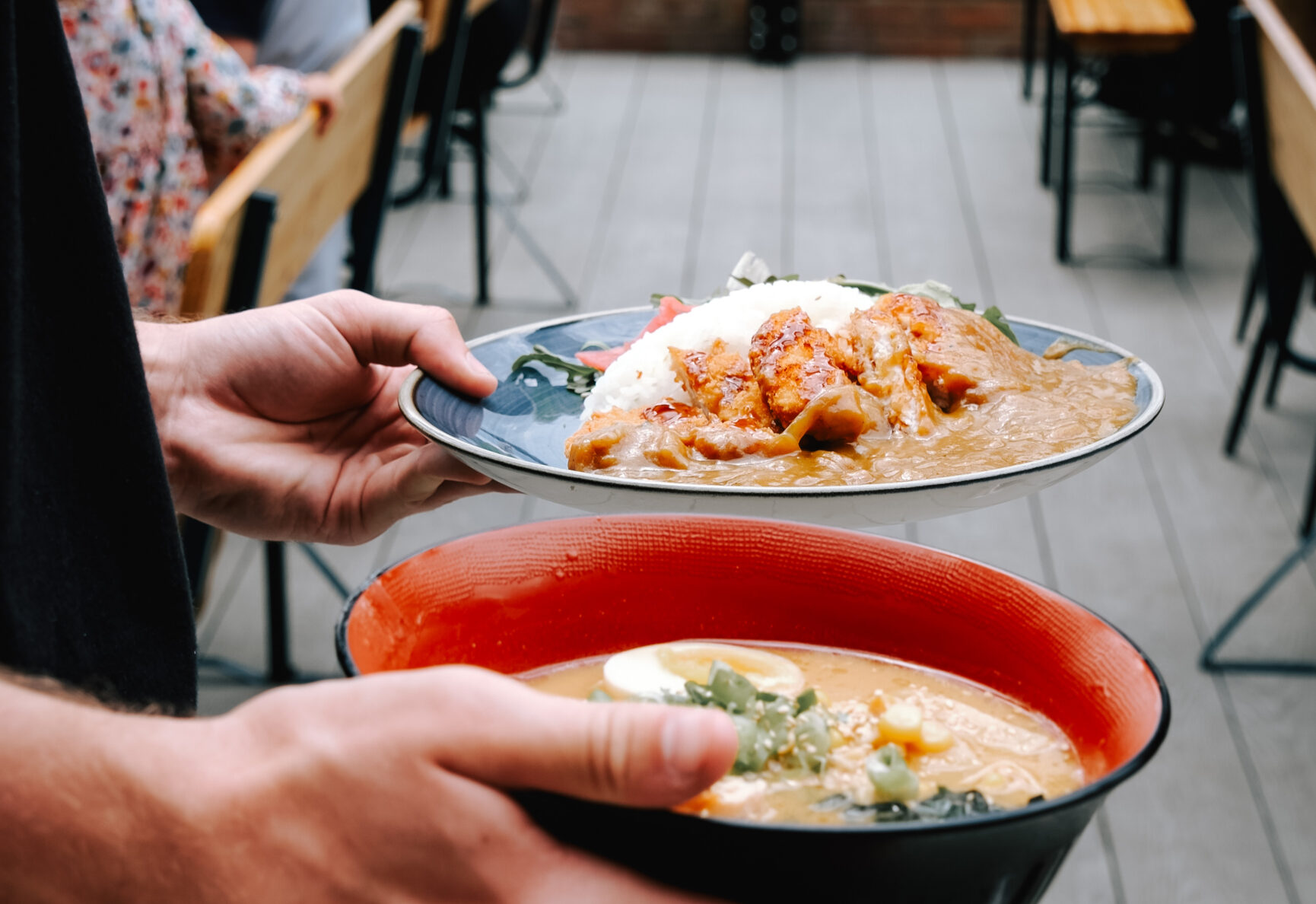 Daikoku Ramen and Katsu Curry being held by a waiters hands. The Food Dock Deck with people sat at tables and chairs can be seen behind, also with a view of the docks water.