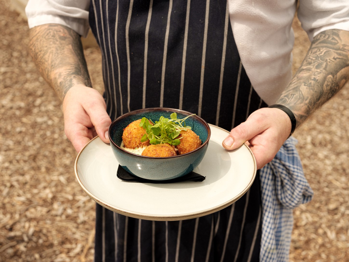 A chef with a dark blue apron with thin, white vertical stripes holds a dish on a serving plate.