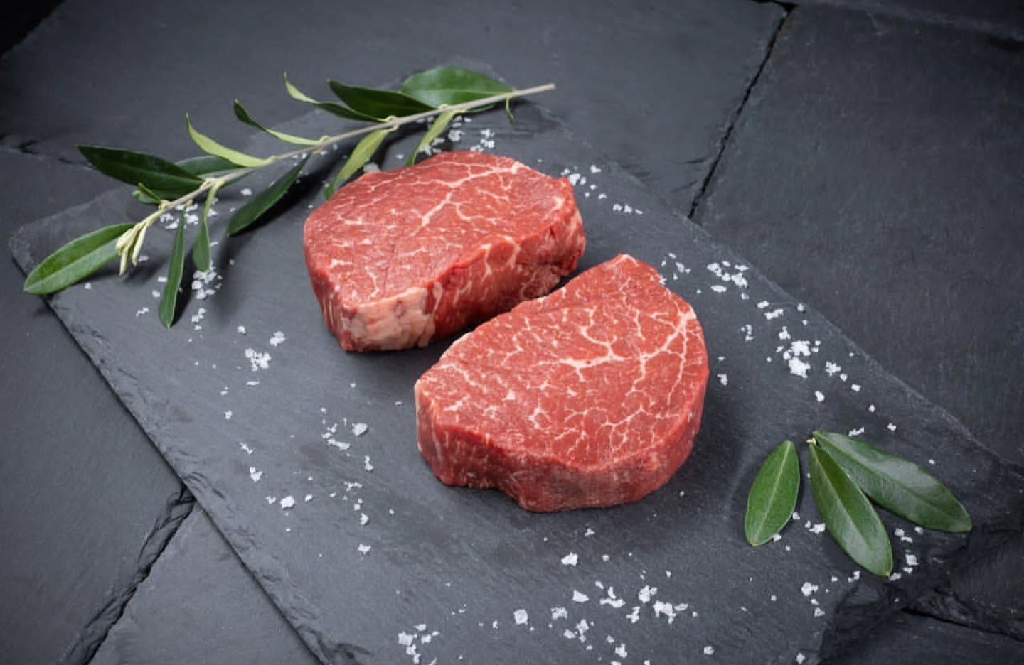 Two British Wagyu steaks on a slate board surrounded by herbs and salt.