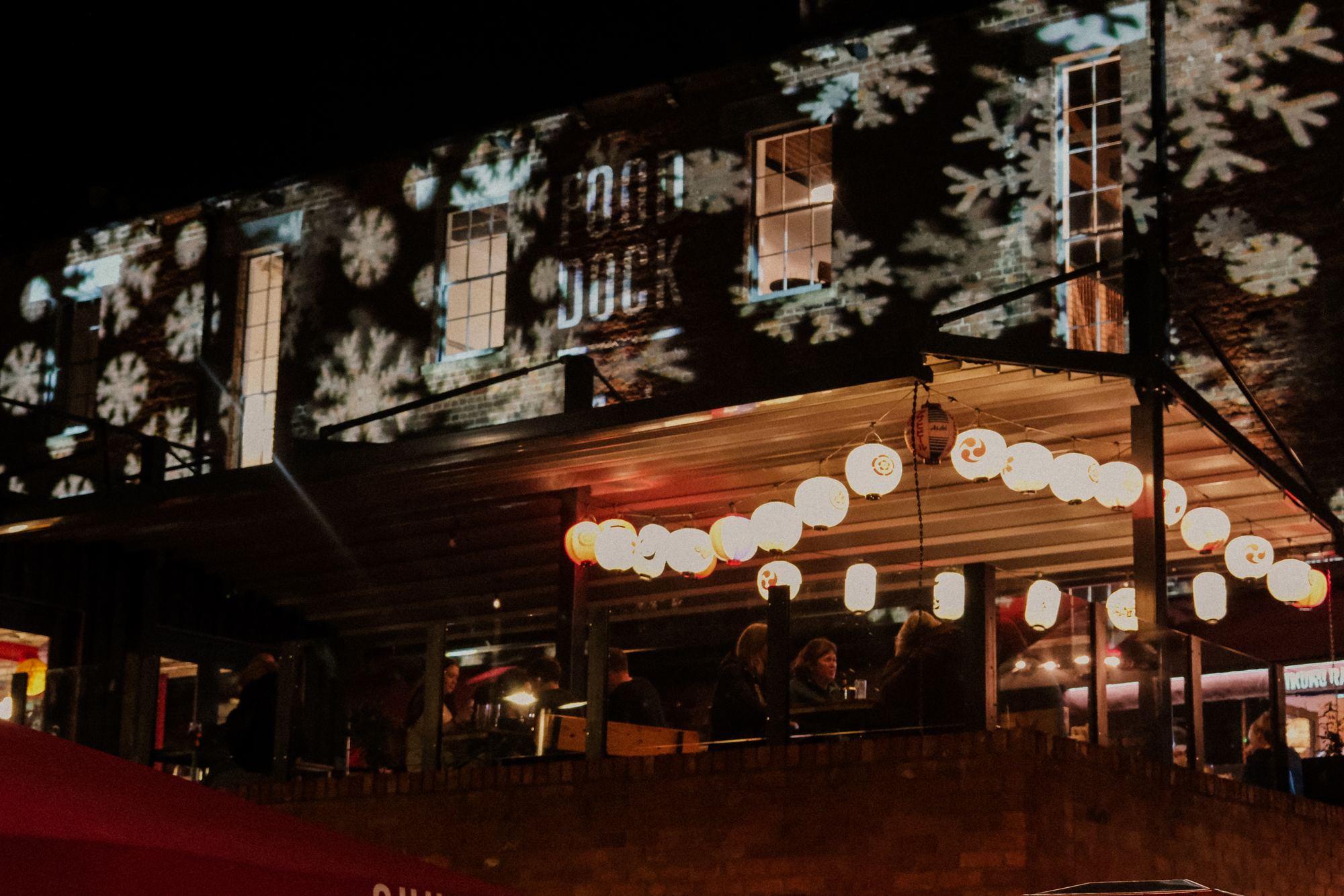 Gloucester Food Dock waterfront terrace at night, strung with paper lanterns and snowflake light projections on the brickwork above